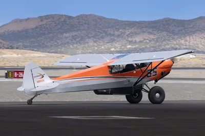 A vibrant orange Carbon Cub aircraft taking off from a runway with a dramatic Montana mountain backdrop.