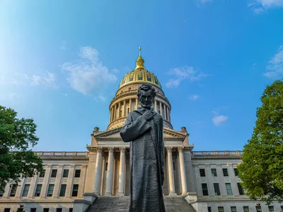 Facade of the West Virginia State Capitol building with a statue of Abraham Lincoln in the foreground under clear blue skies.