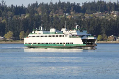 A Washington State Ferry sailing on the calm waters near Port Orchard with a forested Pacific Northwest landscape in the background.