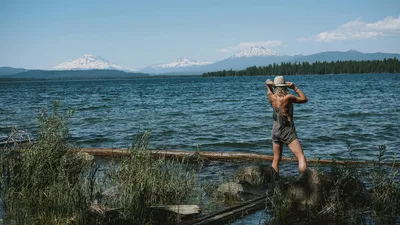 A woman stands by a tranquil lake in Salem, Oregon, with the majestic Cascade Mountains in the background.