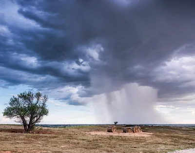 A striking scene of a storm brewing over a plain with hay bales and a lone tree in Oklahoma.